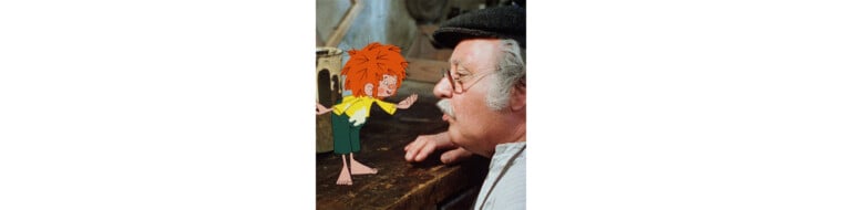 Pumuckl steht auf einer Werkbank und spricht mit dem alten Meister Eder, der ihn neugierig anschaut.